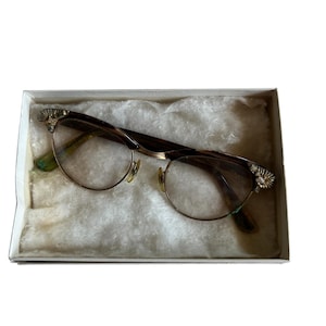 May include: Vintage eyeglasses with a gold-tone metal frame and clear lenses. The glasses have decorative floral accents on the temples and are presented in a white box with a soft, off-white lining. A classic eyewear accessory.