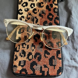 May include: Vintage cat-eye eyeglasses with a silver frame rest on a leopard print eyeglass case. The case is a tapestry of black, brown, and tan. The glasses have clear lenses and a decorative design on the frame.
