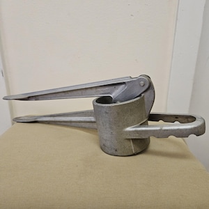 May include: A silver garlic press with a cylindrical chamber and long lever arms. The press has a handle and a hinged design. The metal tool is designed for pressing garlic cloves. The press is made of metal.