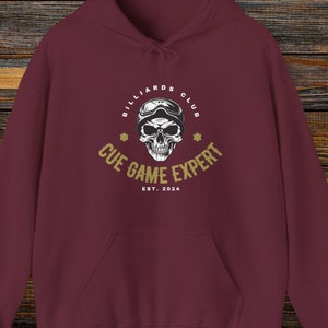 Puede incluir: Sudadera con capucha color burdeos con un logotipo dorado que dice "Billiards Club Cue Game Expert Est. 2024" y una calavera con casco.