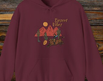 Può includere: Felpa con cappuccio color borgogna con una grafica di un cowboy che cavalca un toro in un paesaggio desertico. Il testo "Desert Vibes" è stampato sulla felpa.