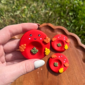 Peut inclure: Trois coupelles rondes en argile rouge ornées d'un visage souriant aux joues roses et de petites feuilles d'automne en relief de couleur orange, jaune et verte. Chaque coupelle présente une indentation centrale pour y déposer de petits objets.