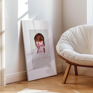 May include: A framed portrait of David Bowie with red hair and a red and blue lightning bolt across his face. The portrait is leaning against a white wall next to a white chair with wooden legs. The image is in a silver frame.