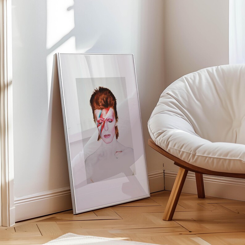 May include: A framed portrait of David Bowie with red hair and a red and blue lightning bolt across his face. The portrait is leaning against a white wall next to a white chair with wooden legs. The image is in a silver frame.