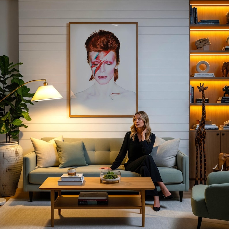 May include: A living room scene featuring a large framed portrait of David Bowie with a red and blue lightning bolt across his face. A woman sits on a light green sofa. A wooden coffee table and a floor lamp are in the foreground.