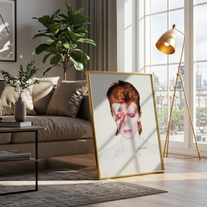 May include: A framed art print featuring a portrait of a person with red hair and a lightning bolt across their face. The artwork is in a gold frame and leans against a wall near a window and a gold floor lamp.