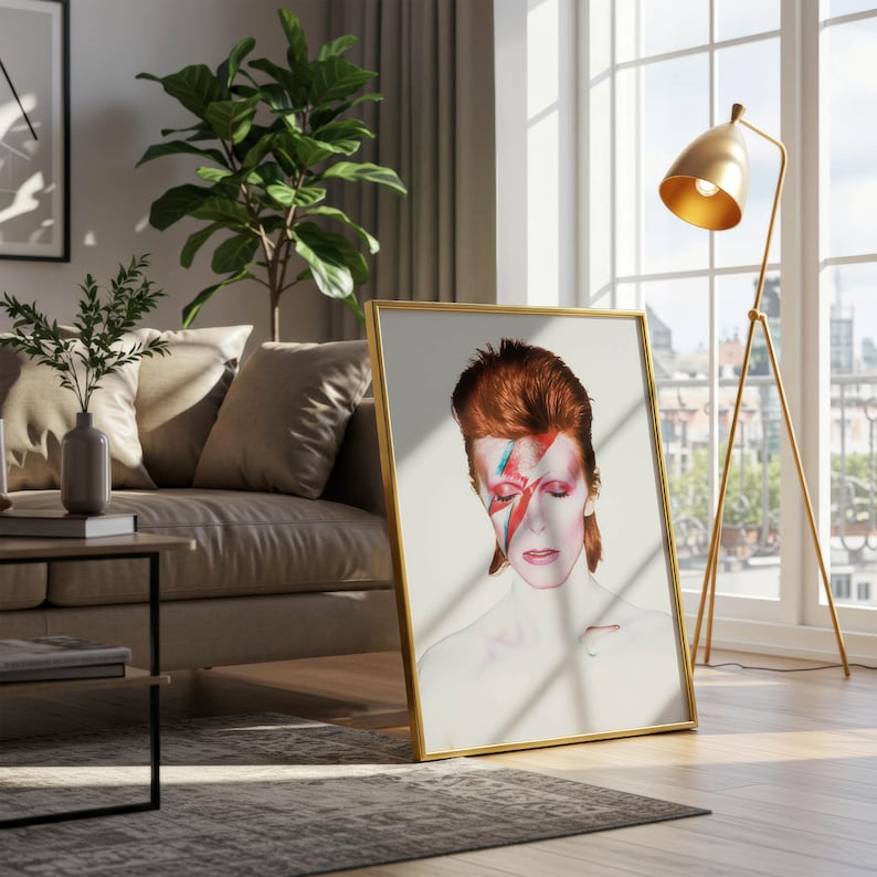 May include: A framed art print featuring a portrait of a person with red hair and a lightning bolt across their face. The artwork is in a gold frame and leans against a wall near a window and a gold floor lamp.