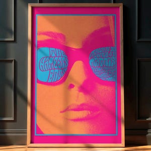 May include: A framed pop art print featuring a close-up of a face wearing large, pink sunglasses with blue lenses. The lenses display the text "THE GREATS ARE BACK" in a stylized font. The background is a gradient of orange and pink.