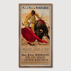 May include: Framed vintage poster advertising a bullfight in Pamplona, Spain. The poster shows a matador in a gold suit with a red cape facing a black bull. Text includes the date, time, and matadors' names. The poster is in a wooden frame.