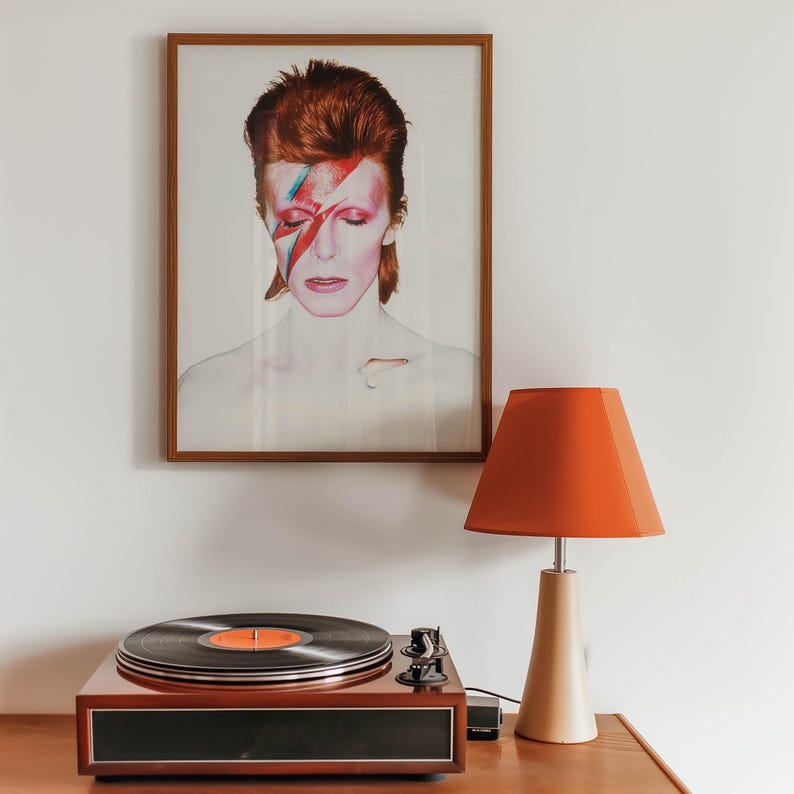 May include: A framed portrait of David Bowie with red hair and a lightning bolt on his face hangs above a record player. An orange lampshade sits atop a beige lamp base. The record player is brown with a black record.