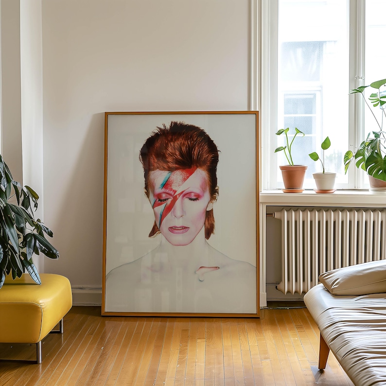 May include: A framed portrait of a person with red hair and a red and blue lightning bolt painted across their face. The portrait is in a room with a yellow ottoman, a white couch, and plants.