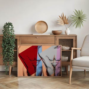 May include: A framed art print featuring a pop art style depiction of a torso. The artwork uses bold colors, including red, blue, and pink, against a neutral background. The print is displayed on a wooden floor, leaning against a wooden cabinet.