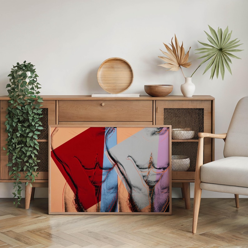 May include: A framed art print featuring a pop art style depiction of a torso. The artwork uses bold colors, including red, blue, and pink, against a neutral background. The print is displayed on a wooden floor, leaning against a wooden cabinet.