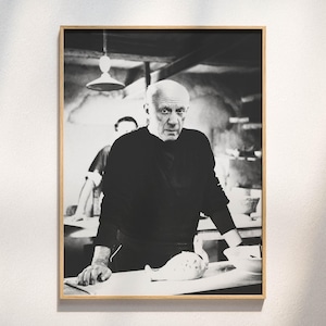 Könnte beinhalten: Ein schwarz-weiß gerahmtes Foto mit einem Porträt von Pablo Picasso in einem dunklen Langarmshirt. Er steht hinter einem Tisch mit einem Keramikobjekt und einer Schale, während im Hintergrund eine Hängelampe zu sehen ist.