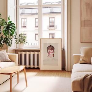 May include: A framed portrait of David Bowie with red and orange hair and a red lightning bolt across his face. The artwork is displayed in a light-filled room with a neutral color palette, a wooden floor, and a large window.