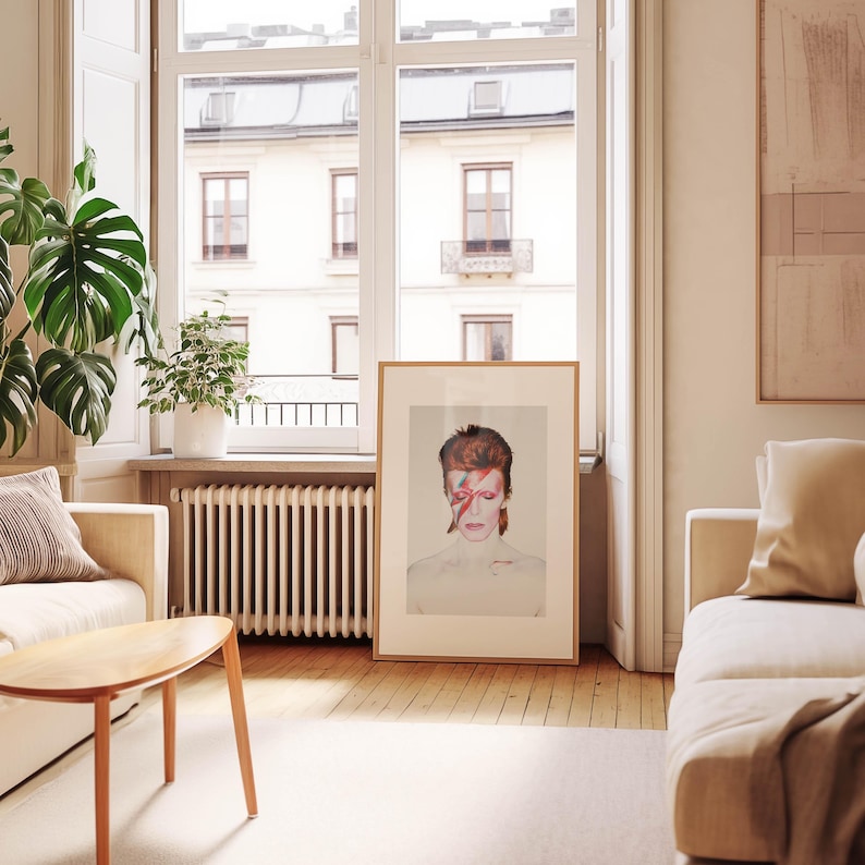 May include: A framed portrait of David Bowie with red and orange hair and a red lightning bolt across his face. The artwork is displayed in a light-filled room with a neutral color palette, a wooden floor, and a large window.