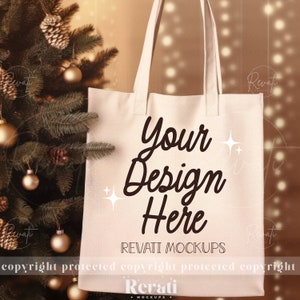 Op de afbeelding: Een crèmekleurige draagtas met de tekst "Your Design Here" in een stijlvol lettertype. De tas staat tegen een achtergrond van een kerstboom versierd met gouden ornamenten en dennenappels. De tekst "REVATI MOCKUPS" is ook zichtbaar.