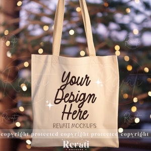 May include: A cream-colored tote bag with the text "Your Design Here" in a dark brown script. The bag has matching cream-colored handles and is set against a blurred background with bokeh lights. The words "REVATI MOCKUPS" are also visible.