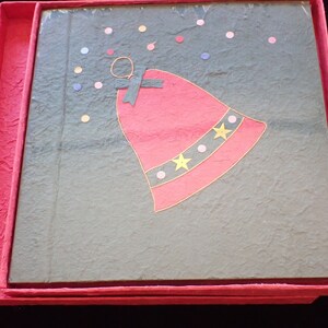 May include: A red and gold bell with a black ribbon hangs from a green background with pink and white polka dots. The bell is in a red box with a textured surface.