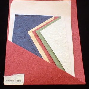 Puede incluir: Una carpeta roja con superficie texturizada contiene una variedad de papel Sa hecho a mano en diferentes colores, incluyendo azul, verde, amarillo y blanco. La carpeta tiene una etiqueta que dice "Handmade Sa Paper".