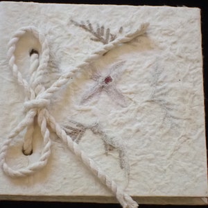 May include: A white handmade journal with a rope closure and pressed flowers and leaves on the cover.