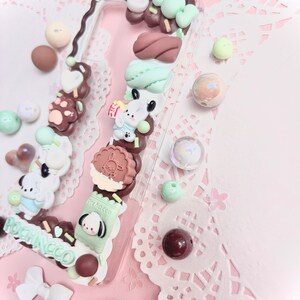 May include: A clear phone case decorated with various pastel-colored charms, including a brown bear paw print, a green and white cookie, and a green and white milk carton with the text "Pocchacco".