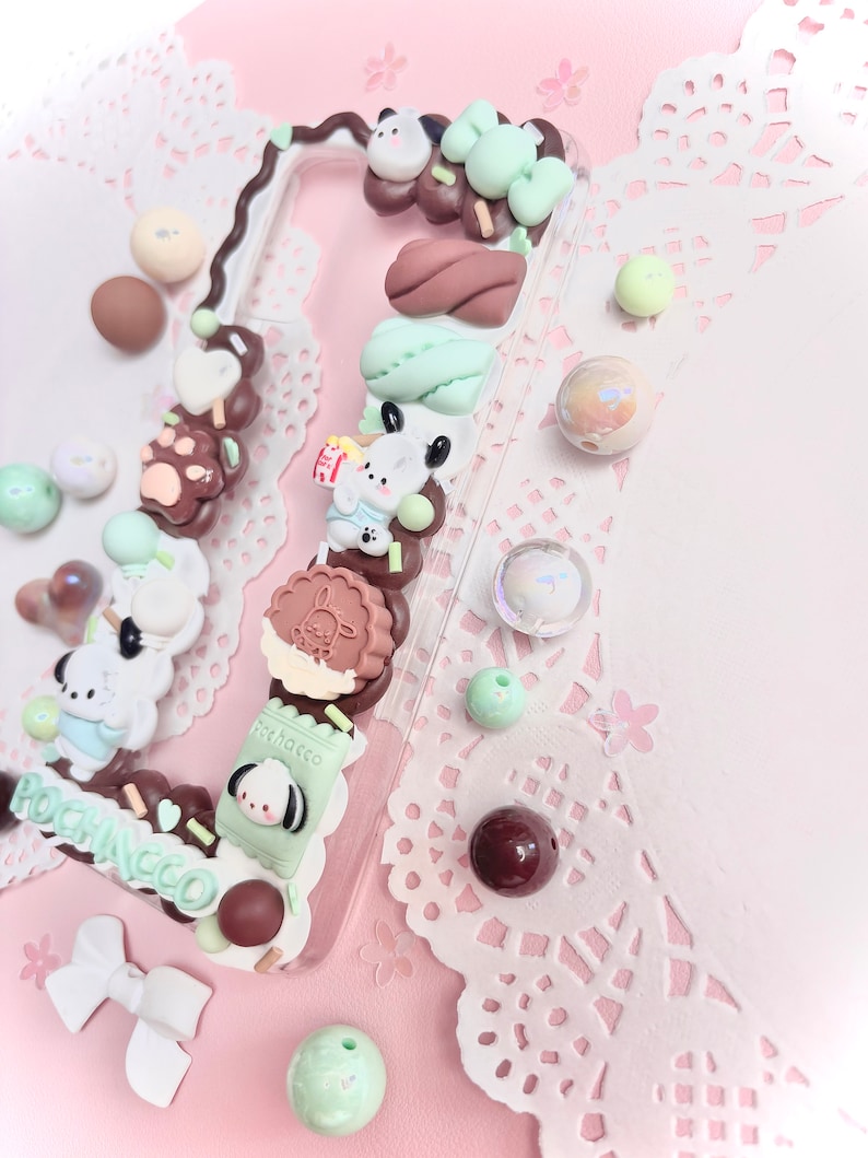 May include: A clear phone case decorated with various pastel-colored charms, including a brown bear paw print, a green and white cookie, and a green and white milk carton with the text "Pocchacco".