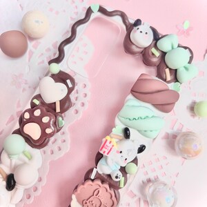 May include: A clear phone case decorated with brown, white, and green 3D embellishments. The embellishments include paw prints, hearts, a dog, and a cookie. The case has a brown swirl design on the side.