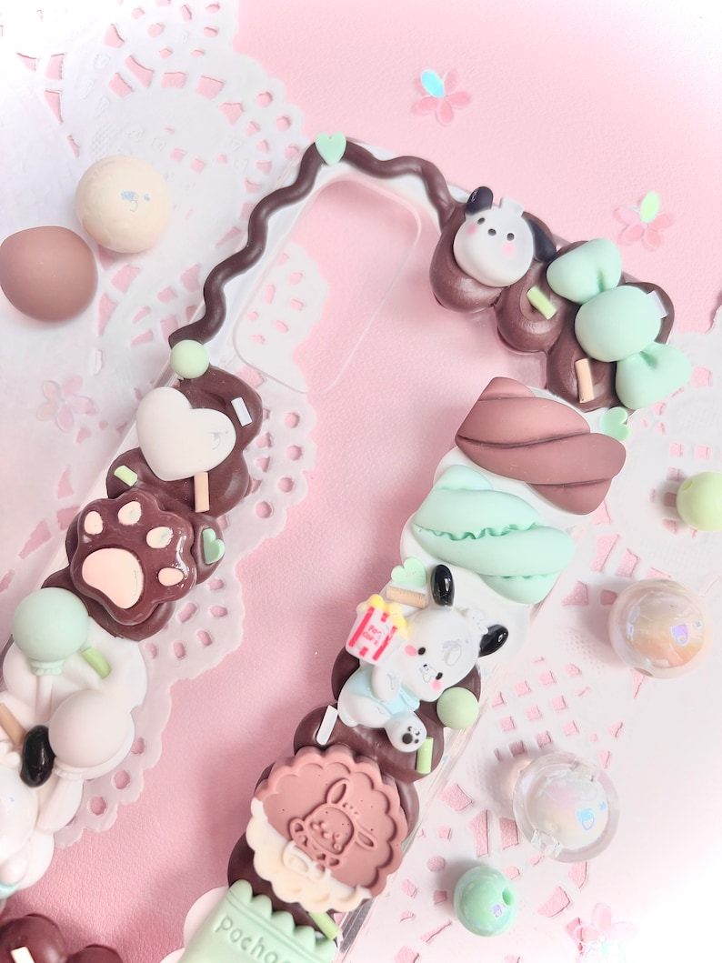 May include: A clear phone case decorated with brown, white, and green 3D embellishments. The embellishments include paw prints, hearts, a dog, and a cookie. The case has a brown swirl design on the side.