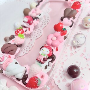 以下が含まれることがあります： ハローキティのキャラクター、イチゴ、チョコレート、その他の可愛い飾りで飾られたピンクの携帯電話ケースです。