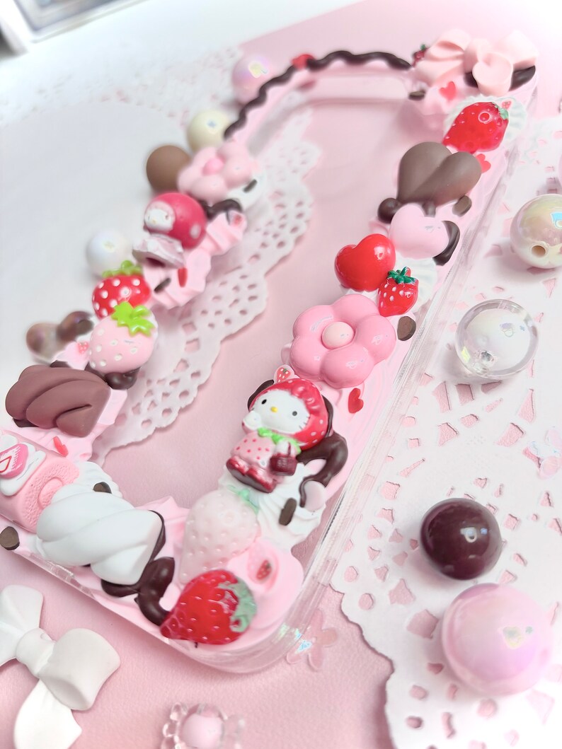 以下が含まれることがあります： ハローキティのキャラクター、イチゴ、チョコレート、その他の可愛い飾りで飾られたピンクの携帯電話ケースです。
