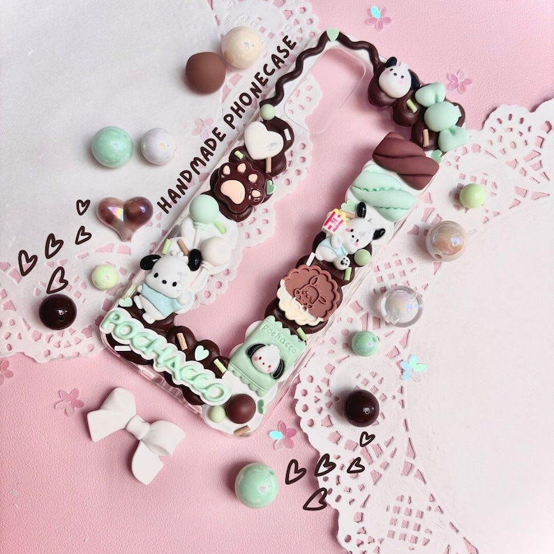 May include: A clear phone case with a brown and white design. The case is decorated with small, colorful charms, including hearts, stars, and a cartoon dog. The text "HANDMADE PHONECASE" is written on the case. The case is surrounded by small, colorful beads and hearts.