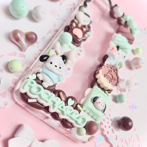 May include: A clear phone case decorated with pastel green, brown, and white 3D charms. The charms include a bear, a dog, a cat, and a variety of other cute designs. The case also features the word "POOHACO" in brown and white lettering.