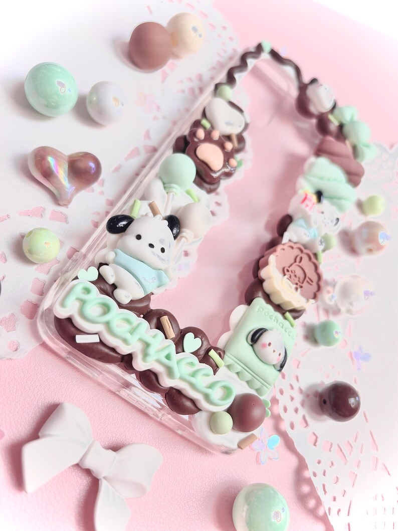 May include: A clear phone case decorated with pastel green, brown, and white 3D charms. The charms include a bear, a dog, a cat, and a variety of other cute designs. The case also features the word "POOHACO" in brown and white lettering.