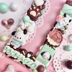 May include: A clear phone case decorated with a variety of colorful charms, including a brown paw print, a white heart, a green balloon, a white dog with a blue bib, a brown cookie with a white bunny, a green box with a white dog, and the word "POCHACCO" in green letters.
