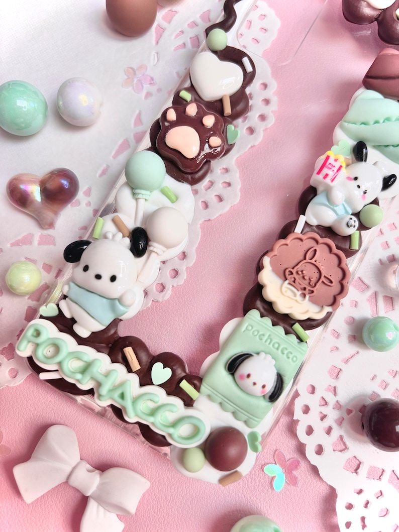 May include: A clear phone case decorated with a variety of colorful charms, including a brown paw print, a white heart, a green balloon, a white dog with a blue bib, a brown cookie with a white bunny, a green box with a white dog, and the word "POCHACCO" in green letters.