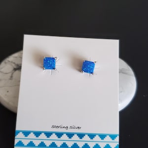 May include: A pair of sterling silver earrings with square blue opal stones. The earrings are displayed on a white card with the text "Sterling Silver" printed on it.
