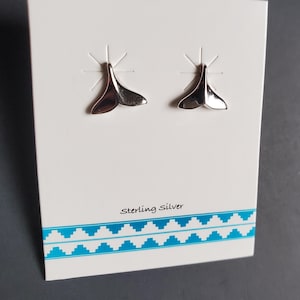 May include: A pair of sterling silver whale tail earrings displayed on a white card with a blue geometric border. The earrings are shiny and reflect light. The card has the words "Sterling Silver" printed on it.