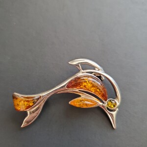 May include: A silver-toned dolphin brooch with amber-colored accents. The brooch features a stylized dolphin design with a curved body and tail, and a small amber stone at the eye. The amber stones are a mix of orange and yellow.