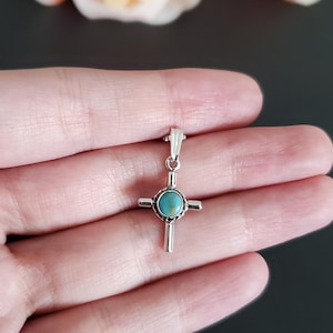 May include: A silver cross pendant with a turquoise stone in the centre. The cross is on a hand against a black background.