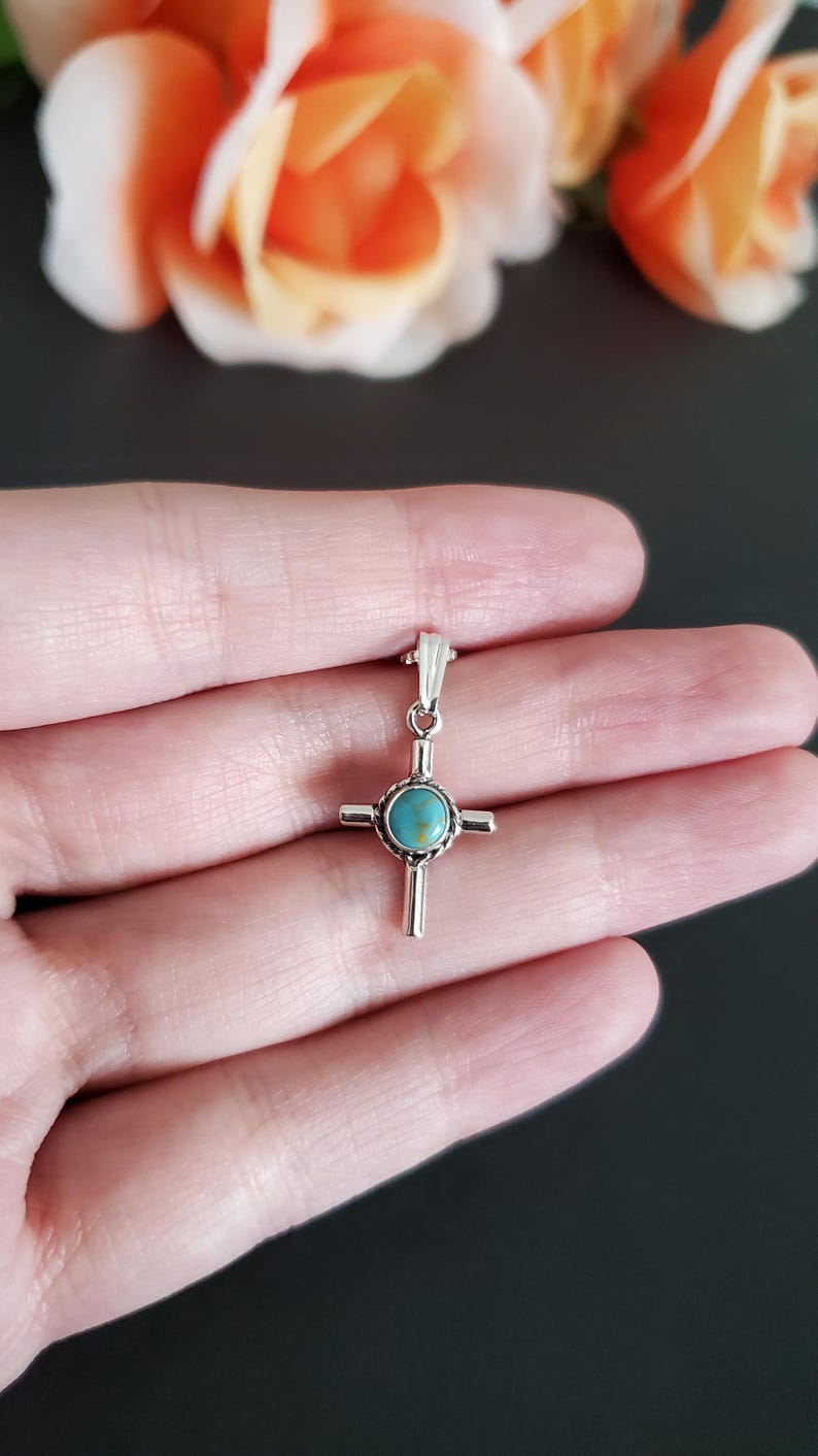 May include: A silver cross pendant with a turquoise stone in the centre. The cross is on a hand against a black background.