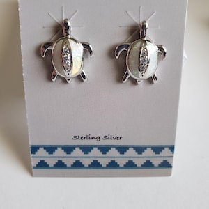 May include: A pair of sterling silver turtle earrings with white opal insets. The earrings are displayed on a white card with a blue and white patterned border. The card also says "Sterling Silver".