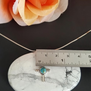 May include: Silver necklace with a turquoise cross pendant. The pendant is approximately 2.5 cm in length. The necklace is on a white marble surface.
