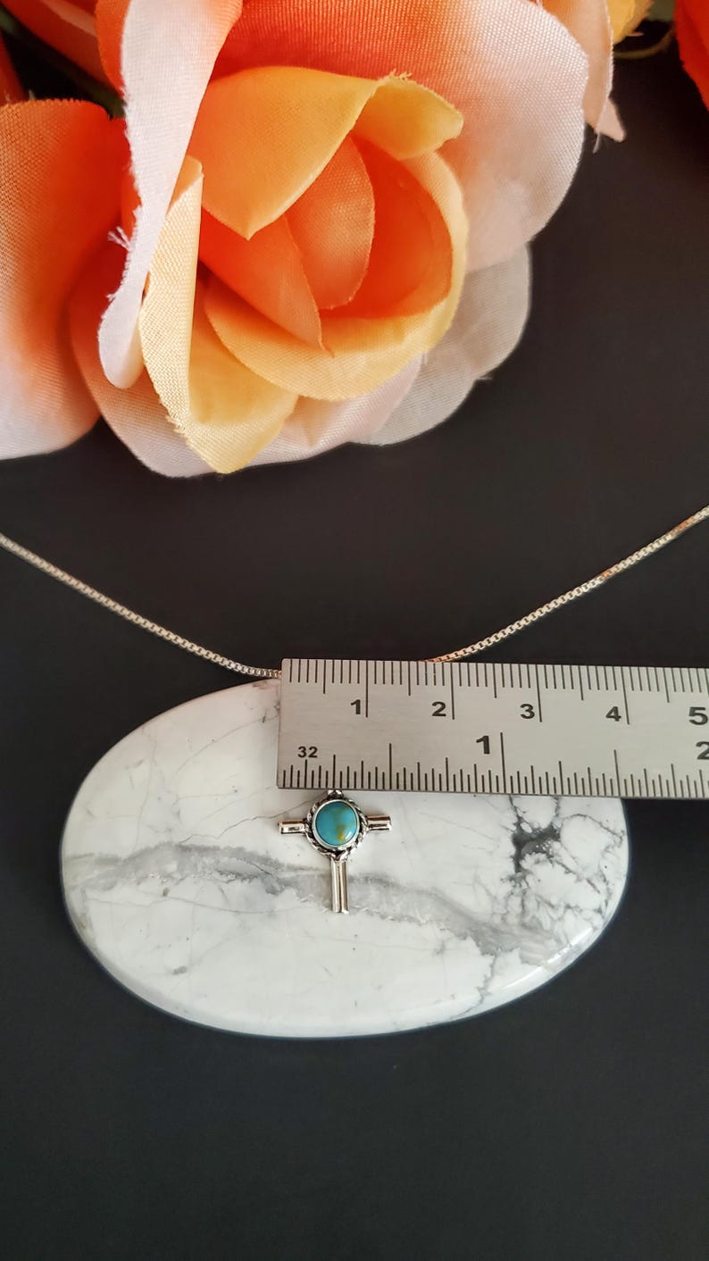 May include: Silver necklace with a turquoise cross pendant. The pendant is approximately 2.5 cm in length. The necklace is on a white marble surface.