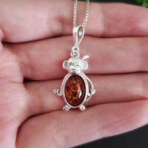 May include: Silver monkey pendant necklace with a polished amber-colored stone. The pendant features a detailed monkey design with a small loop for the chain. The chain is a delicate silver box chain.