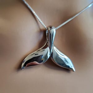 Puede incluir: Un collar de plata con un colgante de cola de ballena. El colgante está hecho de metal pulido y tiene un acabado liso y brillante.