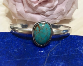 May include: A silver bangle bracelet featuring a turquoise stone set in a silver bezel. The stone is a light blue colour with brown veins.