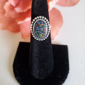 May include: Oval-shaped silver ring with a vibrant, multicolored opal stone. The opal displays flashes of blue, green, and orange. The ring is set on a black velvet display stand, with a soft pink flower in the background.