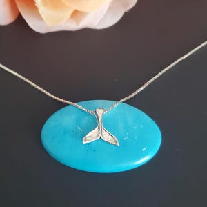 Puede incluir: Un collar de plata con un pequeño colgante en forma de cola de ballena. El collar está sobre una piedra azul turquesa.