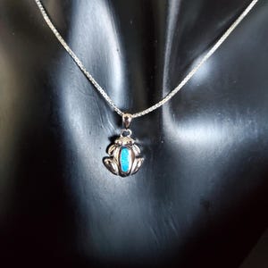 May include: A silver-toned necklace featuring a frog pendant with a turquoise-colored center. The frog pendant is detailed with textured features and is suspended from a delicate chain. The necklace is displayed against a dark background.